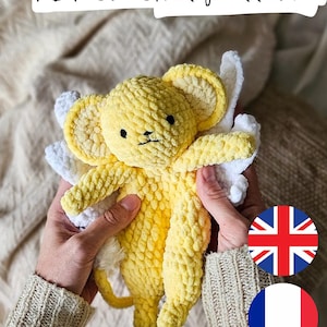 Peut inclure: Une souris jaune au crochet avec des ailes blanches, tenue par une personne. Le texte "PDF crochet pattern" est en haut de l'image. La souris a des yeux noirs et une petite queue. L'image comprend également les drapeaux britannique et français.