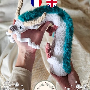 Peut inclure: Un dragon au crochet fait à la main, blanc, turquoise et rose, tenu dans les mains. Le dragon a une texture duveteuse et des détails. Le texte en haut indique "Haku PDF Crochet Pattern".