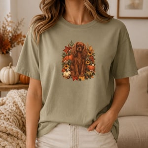 May include: A sage green short-sleeve t-shirt featuring a central graphic of a brown spaniel dog surrounded by autumn elements including orange pumpkins, yellow sunflowers, and colorful fall leaves in shades of red, orange, and gold.