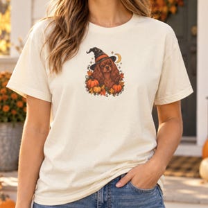 May include: A cream-colored short-sleeve t-shirt featuring a central graphic of a brown spaniel dog wearing a black witch hat, surrounded by orange pumpkins, autumn leaves, and a small yellow crescent moon against a light background.