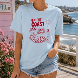 Könnte beinhalten: Hellblaues T-Shirt mit roter Grafik "ON THE COAST". Die Grafik zeigt einen Hund mit herzförmiger Sonnenbrille, einen Sonnenschirm und ein Segelboot. Das T-Shirt hat kurze Ärmel.