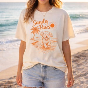 May include: Cream-colored t-shirt with an orange graphic of a puppy running in the ocean, a palm tree, and the words "Sandy Paws." The sun is also depicted in the graphic. The shirt is paired with denim shorts.