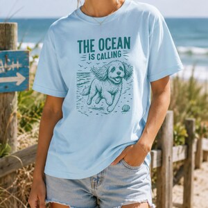 May include: Light blue t-shirt with the text "THE OCEAN IS CALLING" and a graphic of a dog running on a beach. The design is in teal. The shirt is a crew neck and has short sleeves.