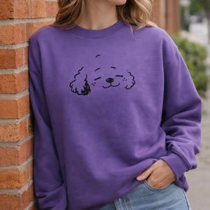 May include: A purple sweatshirt with a black cartoon dog face. The dog has closed eyes and a smiling mouth. The sweatshirt has a crew neck and long sleeves.