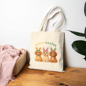 May include: A cream-colored tote bag featuring three adorable puppies wearing Easter bunny ears. The puppies are in shades of brown and cream, with the words "HAPPY EASTER" in pastel colors above them. The bag has a simple, clean design.