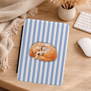 May include: A tablet case with a blue and white striped pattern. The case features a sleeping, fluffy, orange-brown dog illustration with the text "LIVING MY BEST LIFE" in white letters. The case is on a wooden surface.