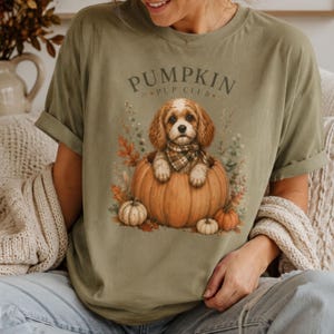 May include: An olive green t-shirt featuring a graphic of a brown and white puppy wearing a plaid scarf, sitting inside a large orange pumpkin. The text PUMPKIN PUP CLUB is printed in an arch above the puppy, surrounded by autumn leaves.