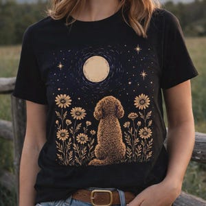 Puede incluir: Camiseta negra con una ilustración de un perro mirando una luna llena en un cielo estrellado, rodeado de flores. El diseño es de estilo caprichoso. La camiseta está hecha de tela suave.