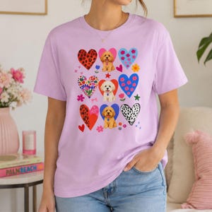 Op de afbeelding: Een lichtpaars t-shirt met een ontwerp van harten en puppy's. De harten hebben verschillende patronen, waaronder luipaardprint, bloemen en stippen. De puppy's worden getoond in verschillende kleuren en rassen, wat een speels element toevoegt.
