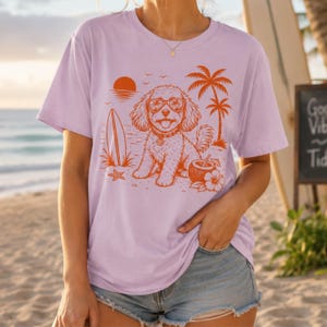 May include: A light purple t-shirt featuring an orange graphic print of a dog wearing sunglasses, sitting on a beach with a surfboard, palm trees, a coconut drink, and a sunset. The dog has a happy expression with its tongue out.