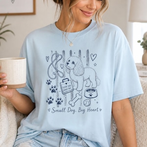 Könnte beinhalten: Hellblaues T-Shirt mit einem Hunde-Motiv. Das Design umfasst eine Hundeillustration, Pfotenabdrücke, eine Leine, einen Hundenapf, einen Knochen und den Text "Small Dog. Big Heart."