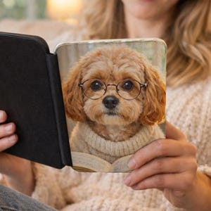 Könnte beinhalten: Eine schwarze E-Reader-Hülle mit einem aufgedruckten Bild eines Cavalier King Charles Spaniel mit runder Brille und einem Pullover. Der Hund schaut auf ein offenes Buch. Die Hülle wird offen gehalten.