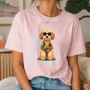 May include: A light pink short-sleeved t-shirt featuring a central graphic of a tan-coloured dog wearing black sunglasses and a colourful tropical floral print shirt. The dog is depicted in a seated position, centred on the chest of the garment.