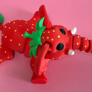 May include: A whimsical red elephant figurine with yellow polka dots, resembling a strawberry. The elephant has green leaves on its head and tail, black eyes, and white tusks. The background is a solid pink.