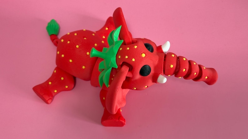 May include: A whimsical red elephant figurine with yellow polka dots, resembling a strawberry. The elephant has green leaves on its head and tail, black eyes, and white tusks. The background is a solid pink.