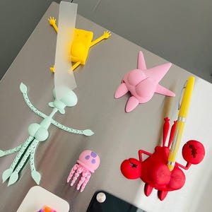 May include: Five 3D printed character magnets in the shape of SpongeBob SquarePants, Patrick Star, Squidward Tentacles, a jellyfish, and a crab. The magnets are yellow, pink, light blue, pink, and red. A yellow pen is stuck in the crab magnet.