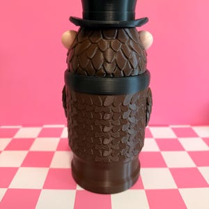 May include: A brown, textured figurine of a character wearing a black top hat and a black band around its neck. The figurine has two small, light-colored spheres on either side of its head. The background is a pink wall and a pink and white checkered surface.