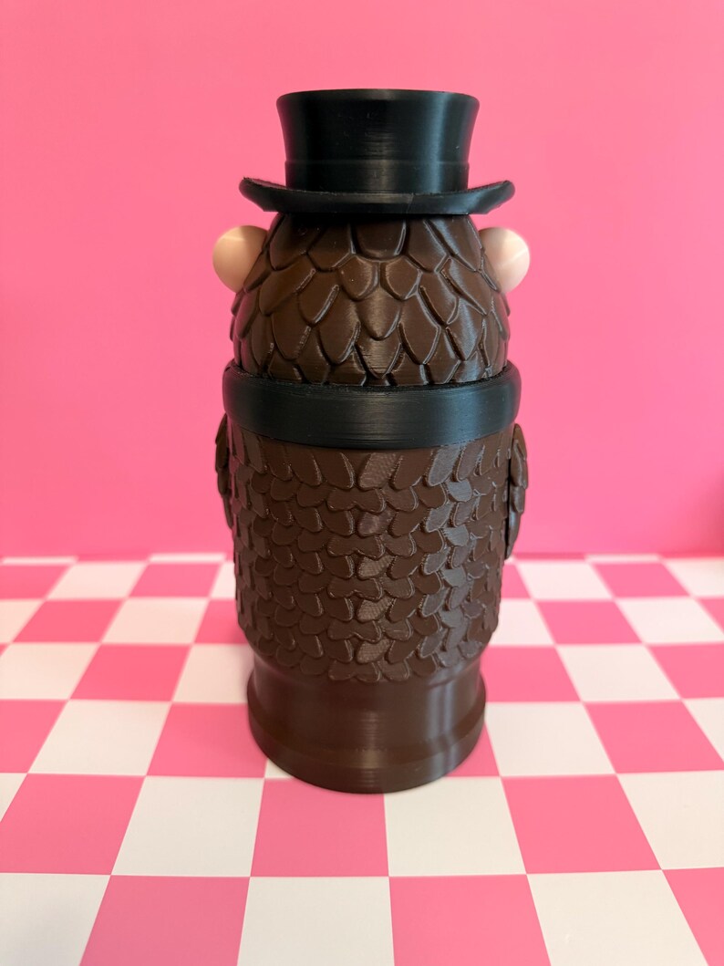May include: A brown, textured figurine of a character wearing a black top hat and a black band around its neck. The figurine has two small, light-colored spheres on either side of its head. The background is a pink wall and a pink and white checkered surface.