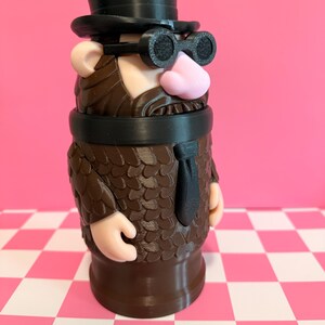 May include: A whimsical, 3D-printed figurine of a character with a top hat, goggles, and a suit. The figure has a pink nose, tan ears, and a brown beard and suit. The base is brown, and the background is pink.
