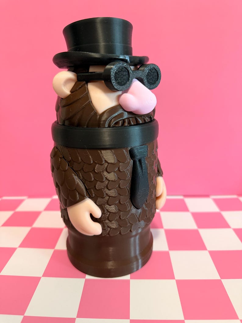 May include: A whimsical, 3D-printed figurine of a character with a top hat, goggles, and a suit. The figure has a pink nose, tan ears, and a brown beard and suit. The base is brown, and the background is pink.