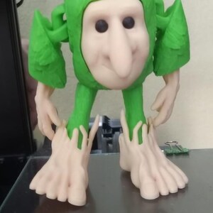 Op de afbeelding: Een grillig 3D-geprint beeldje van een wezen met een lichtgekleurd gezicht, zwarte ogen en een lange neus. Het draagt een groene outfit met capuchon en bladvormige accenten en heeft grote voeten. Het beeldje staat op een donkere ondergrond.