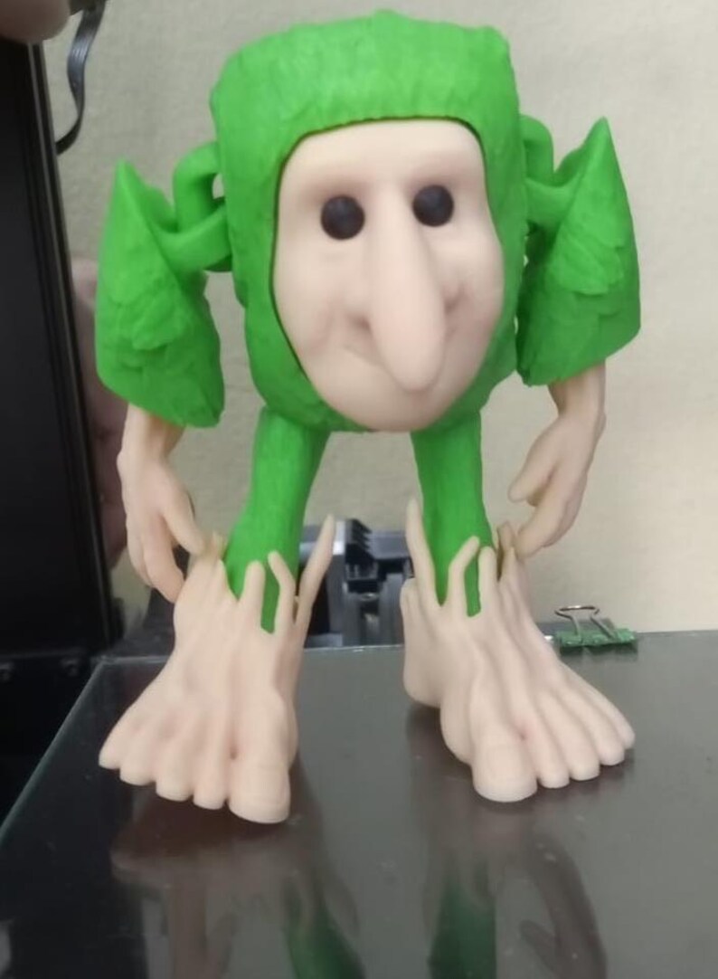 Op de afbeelding: Een grillig 3D-geprint beeldje van een wezen met een lichtgekleurd gezicht, zwarte ogen en een lange neus. Het draagt een groene outfit met capuchon en bladvormige accenten en heeft grote voeten. Het beeldje staat op een donkere ondergrond.