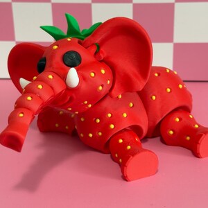 May include: A whimsical red elephant sculpture with yellow dots, resembling a strawberry. The elephant has large ears, a trunk, and green leaves on its head. The sculpture is on a pink surface.
