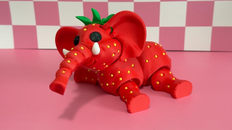 May include: A whimsical red elephant sculpture with yellow dots, resembling a strawberry. The elephant has large ears, a trunk, and green leaves on its head. The sculpture is on a pink surface.