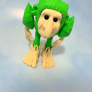 Op de afbeelding: Een grillige groene figuur met een wit gezicht, lange neus en zwarte ogen. De figuur heeft bladachtige aanhangsels en grote voeten. De figuur staat op een lichtblauwe achtergrond.