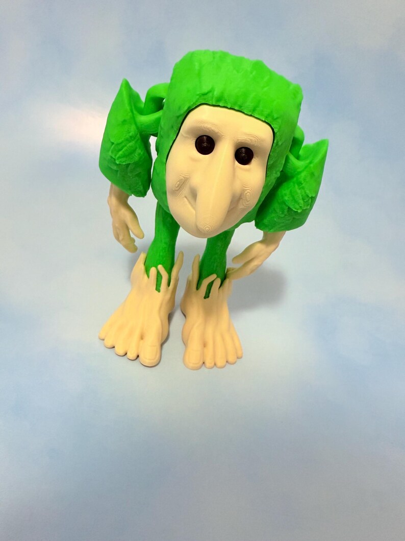 Op de afbeelding: Een grillige groene figuur met een wit gezicht, lange neus en zwarte ogen. De figuur heeft bladachtige aanhangsels en grote voeten. De figuur staat op een lichtblauwe achtergrond.