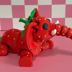 May include: A whimsical red elephant figurine with a strawberry design. The elephant has yellow seeds, green leaves, and white tusks. The trunk is segmented. The figurine is on a pink surface.