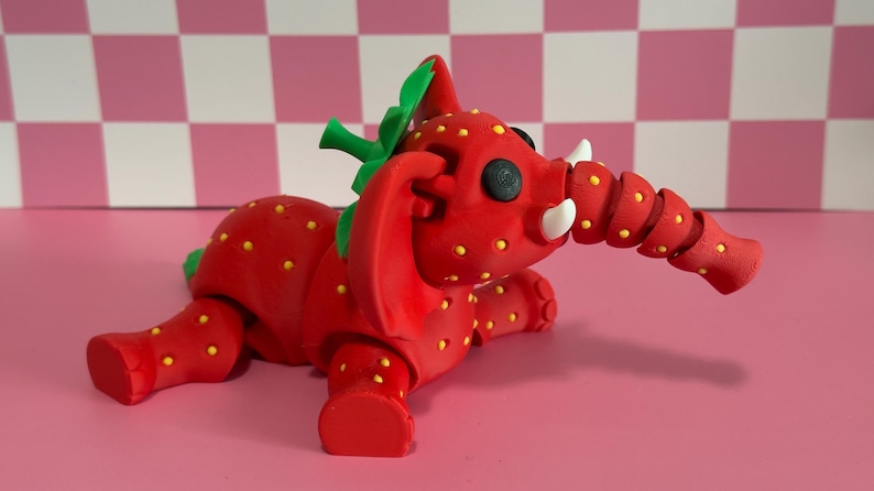 May include: A whimsical red elephant figurine with a strawberry design. The elephant has yellow seeds, green leaves, and white tusks. The trunk is segmented. The figurine is on a pink surface.