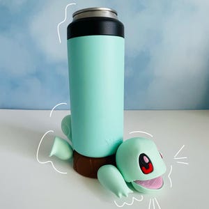 Peut inclure: Une bouteille d'eau réutilisable vert clair avec un couvercle noir et une base brune. La bouteille est tenue par une tortue de dessin animé verte et rouge.