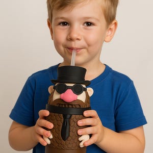May include: A child in a blue shirt is drinking from a novelty tumbler. The tumbler is brown with a cartoon face, black top hat, sunglasses, and a pink nose. The tumbler has a black tie and a clear straw.