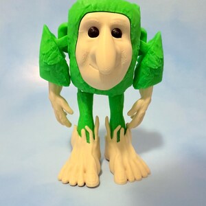 Op de afbeelding: Een grillig groen beeldje met een lichtbeige gezicht, handen en voeten. De figuur heeft een vierkant hoofd, een grote neus en zwarte ogen. De armen en benen zijn dun en de voeten zijn groot en gespreid. De achtergrond is lichtblauw.