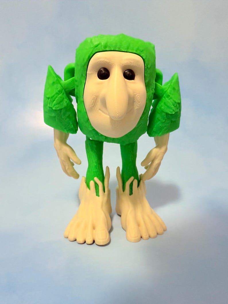 Op de afbeelding: Een grillig groen beeldje met een lichtbeige gezicht, handen en voeten. De figuur heeft een vierkant hoofd, een grote neus en zwarte ogen. De armen en benen zijn dun en de voeten zijn groot en gespreid. De achtergrond is lichtblauw.