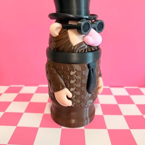 May include: A whimsical, 3D-printed figurine of a man with a top hat, round glasses, and a beard. The figure has a pink nose and ears, a black tie, and a brown textured coat. The background is pink with a checkered pink and white surface.