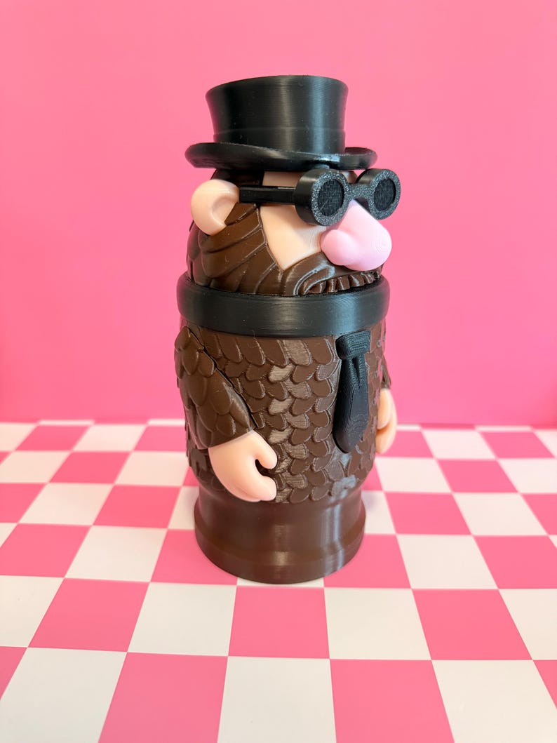 May include: A whimsical, 3D-printed figurine of a man with a top hat, round glasses, and a beard. The figure has a pink nose and ears, a black tie, and a brown textured coat. The background is pink with a checkered pink and white surface.