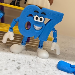 Puede incluir: Una figura azul impresa en 3D con forma de número 67, con cara sonriente, cejas negras y ojos blancos. El personaje tiene brazos y piernas azules articulados con manos y zapatos blancos. Una pieza de extremidad azul articulada yace cerca.