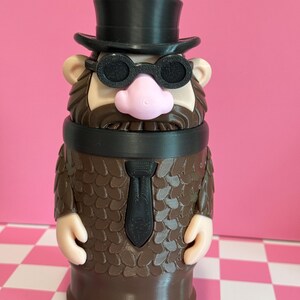 May include: A novelty tumbler shaped like a cartoon character with a black top hat, glasses, and tie. The brown body has a textured pattern, with a pink nose and tan ears. A purple straw extends from the top.