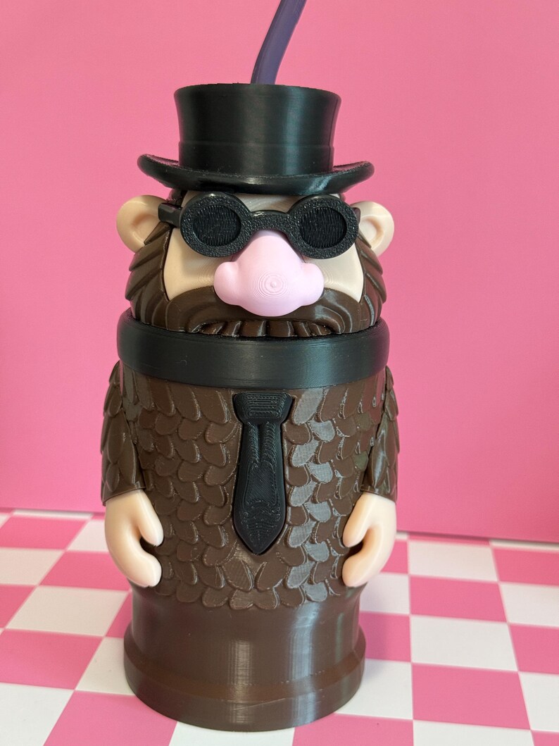 May include: A novelty tumbler shaped like a cartoon character with a black top hat, glasses, and tie. The brown body has a textured pattern, with a pink nose and tan ears. A purple straw extends from the top.