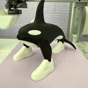 May include: A 3D-printed orca figurine in black and white, wearing white trainers. The orca has a dorsal fin and white patches. The figurine is placed on a light purple surface, with a 3D printer in the background.