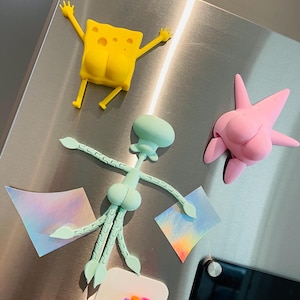May include: A set of three 3D printed magnets featuring characters from the cartoon Spongebob Squarepants. The magnets are yellow, pink, and light blue. The yellow magnet is Spongebob, the pink magnet is Patrick Star, and the light blue magnet is Squidward Tentacles. The magnets are attached to a stainless steel refrigerator.