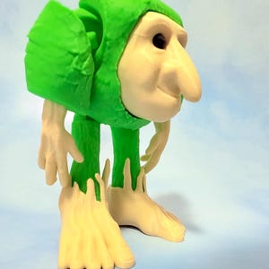 Op de afbeelding: Een grillig beeldje van een trollachtig wezen. Het heeft een grote, bolle neus, kleine zwarte ogen en een groene outfit met capuchon. De handen en voeten zijn lichtgeel, met langwerpige vingers en tenen. Het figuur staat in een licht gebogen houding.