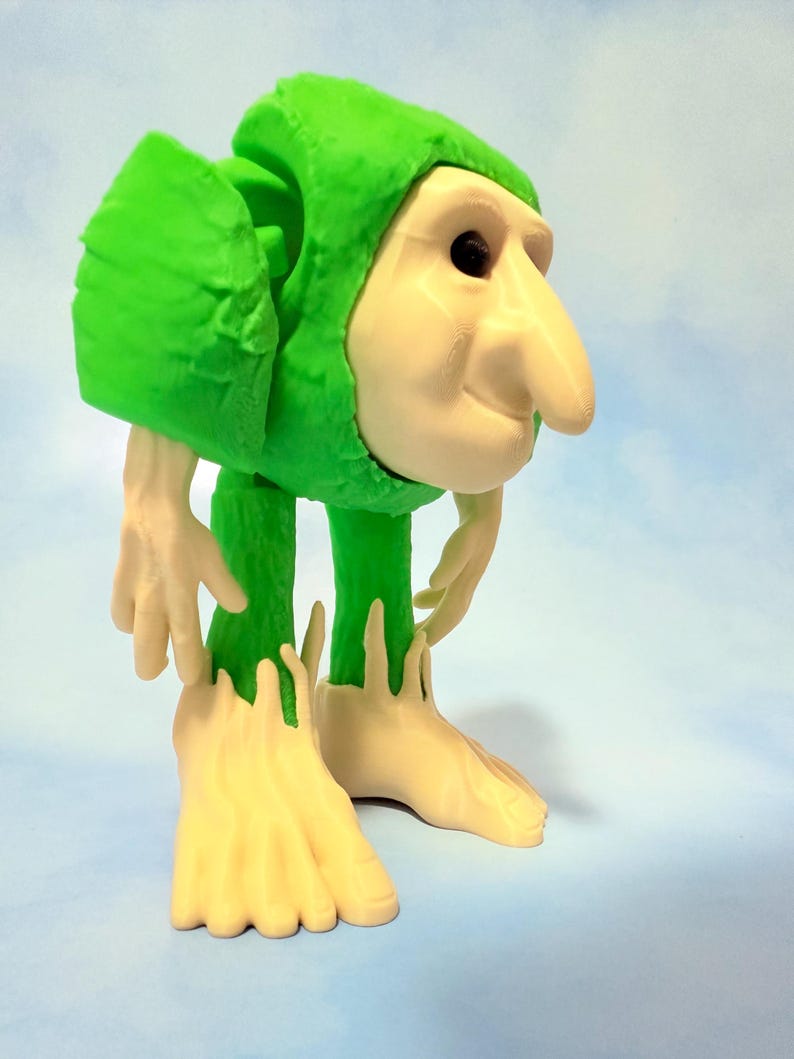 Op de afbeelding: Een grillig beeldje van een trollachtig wezen. Het heeft een grote, bolle neus, kleine zwarte ogen en een groene outfit met capuchon. De handen en voeten zijn lichtgeel, met langwerpige vingers en tenen. Het figuur staat in een licht gebogen houding.
