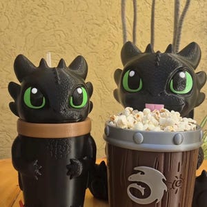 May include: Two black character-themed containers. One is a cup with a brown band, the other is a popcorn bucket. Both have green-eyed faces. The popcorn bucket is filled with popcorn and has a silver emblem.