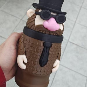 May include: A novelty tumbler shaped like a cartoon character with a black top hat, glasses, and tie. The tumbler is brown with a pink nose and a straw. The design resembles a distinguished gentleman.