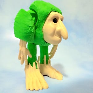 Op de afbeelding: Een grillig beeldje van een wezen met een groene hoodie en lichtbruin gezicht, handen en voeten. De figuur heeft een lange neus en staat in een gebogen houding. De achtergrond is een lichtblauwe gradi&euml;nt.