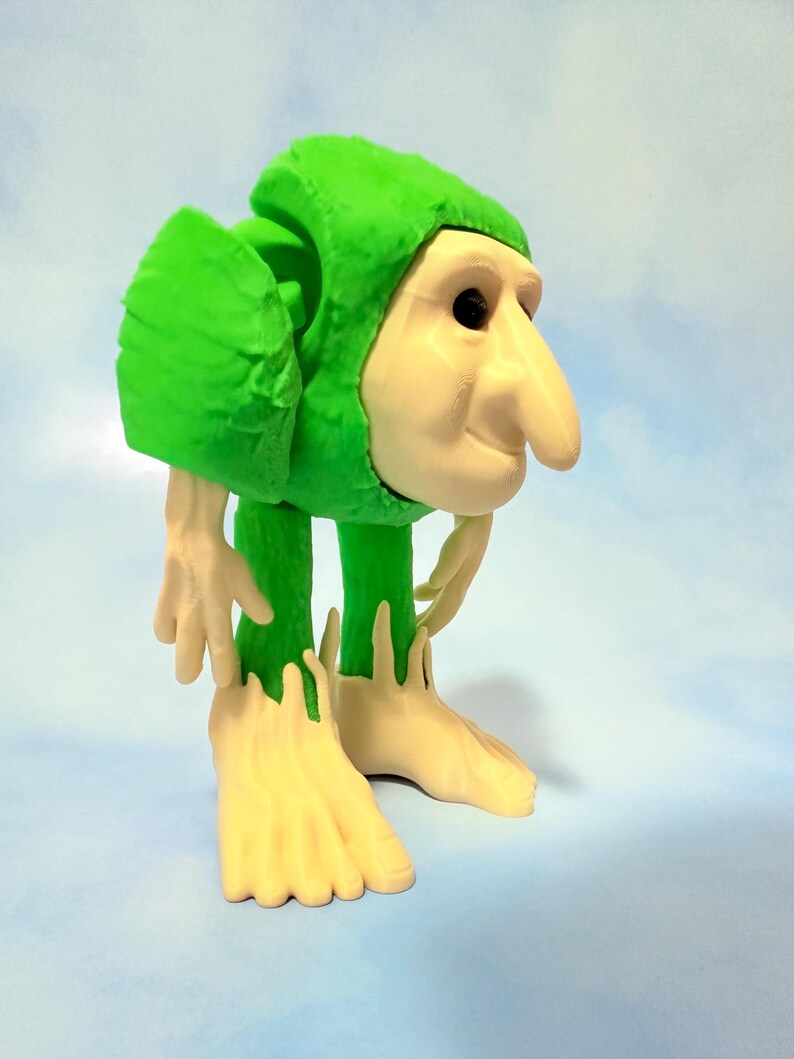 Op de afbeelding: Een grillig beeldje van een wezen met een groene hoodie en lichtbruin gezicht, handen en voeten. De figuur heeft een lange neus en staat in een gebogen houding. De achtergrond is een lichtblauwe gradi&euml;nt.