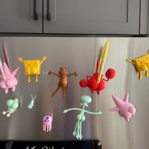 Puede incluir: Una colección de imanes de personajes de dibujos animados coloridos, incluyendo Bob Esponja y otros personajes, se exhiben en un refrigerador de acero inoxidable. Los imanes son de varios colores, incluyendo amarillo, rosa, rojo y verde.